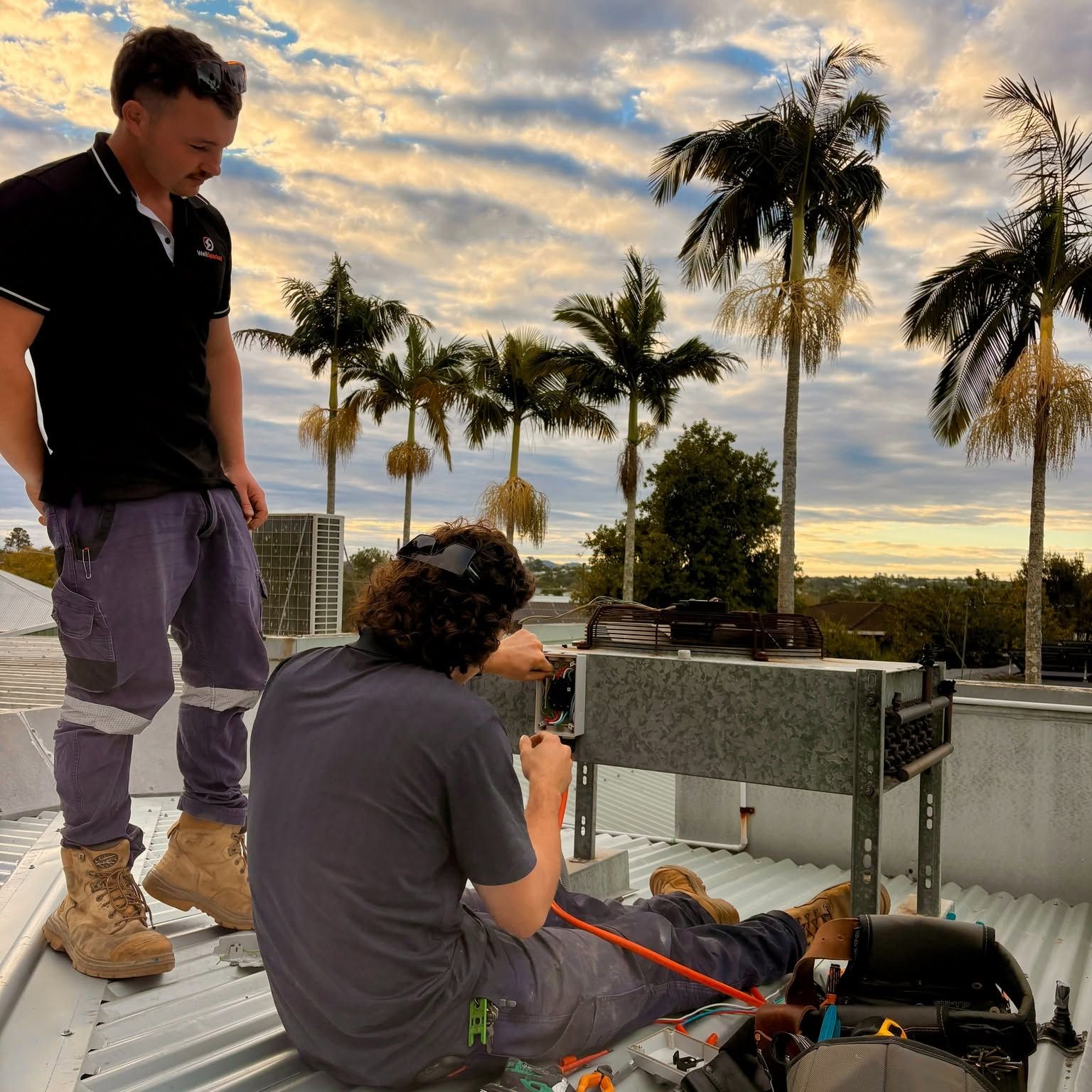 Rory standing on a roof at sunset while Oscar wires an outdoor unit