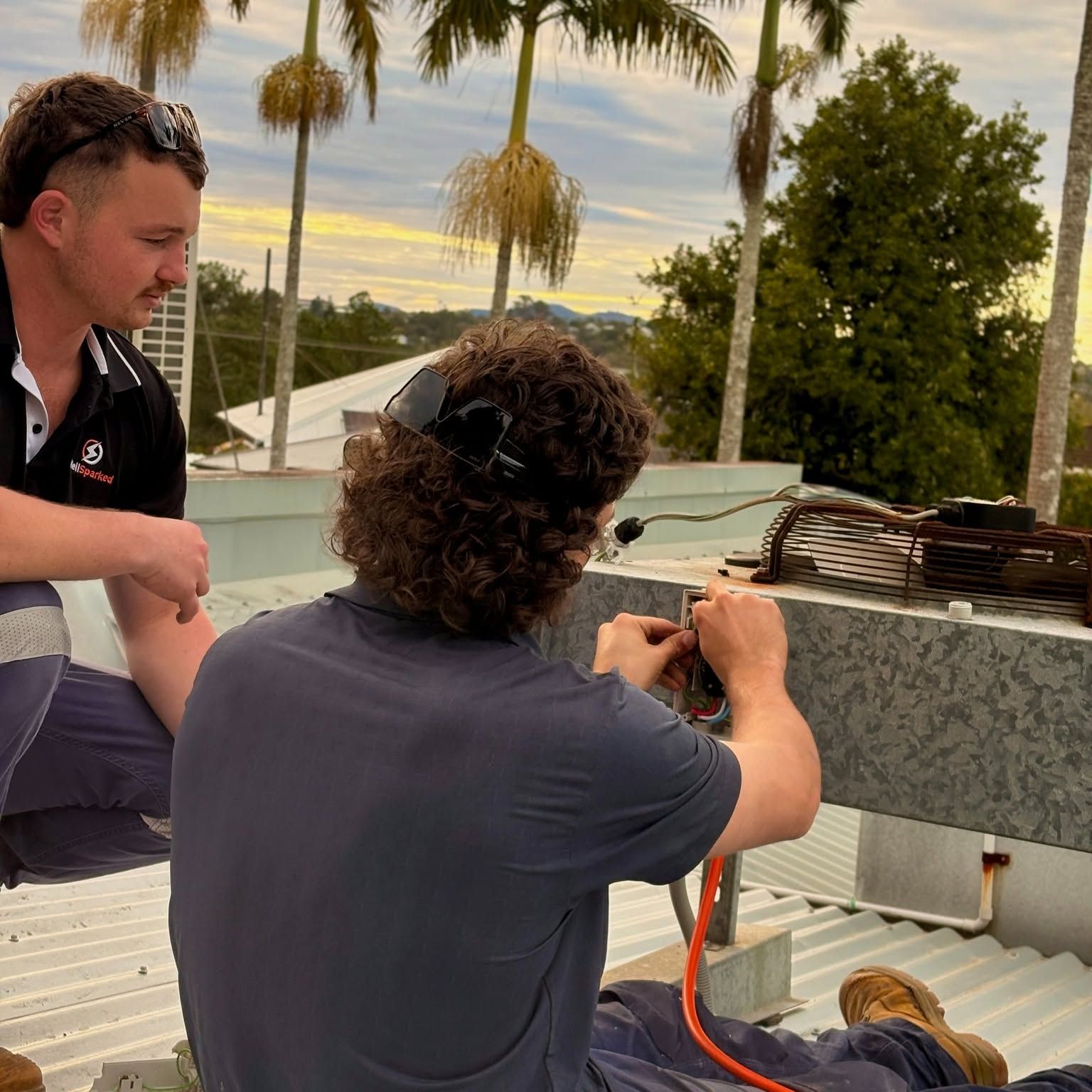 Oscar connecting wiring to a rooftop unit while Rory looks on